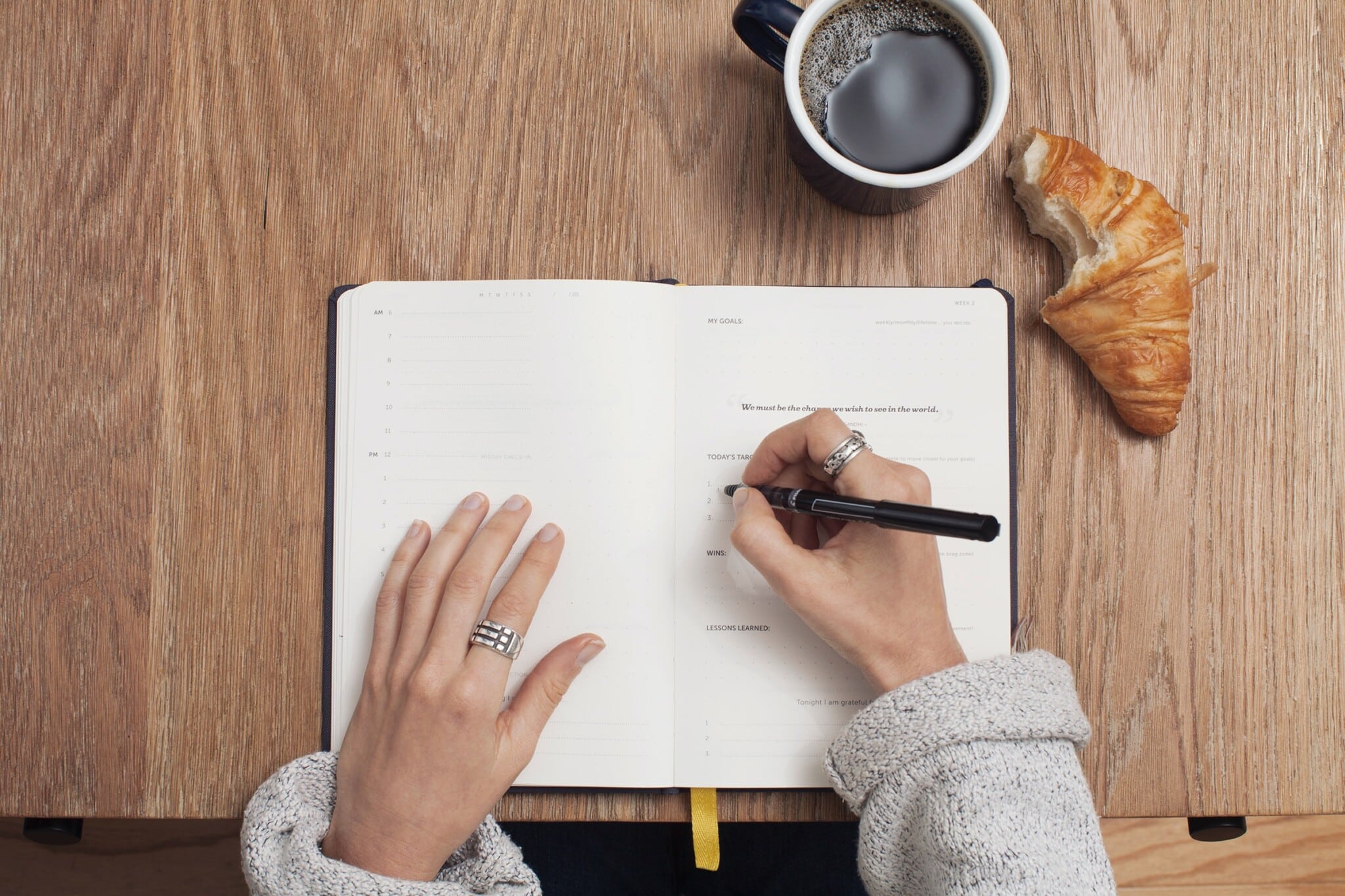 Bird's-eye view of a person writing in a journal next to a cup of coffee and a half-eaten croissant.